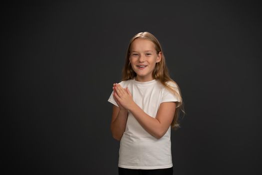 Excited little girl inspired by parents with hands put together, happily looking at the camera wearing white t-shirt and black pants isolated on black background.