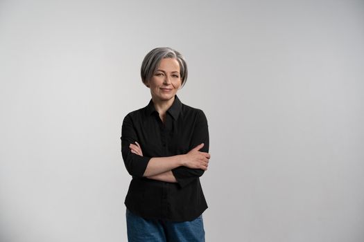 Pretty mid aged grey haired woman in black shirt isolated on grey background. Confident mature grey haired woman looking on camera with arms folded. Human emotions, facial expression concept.