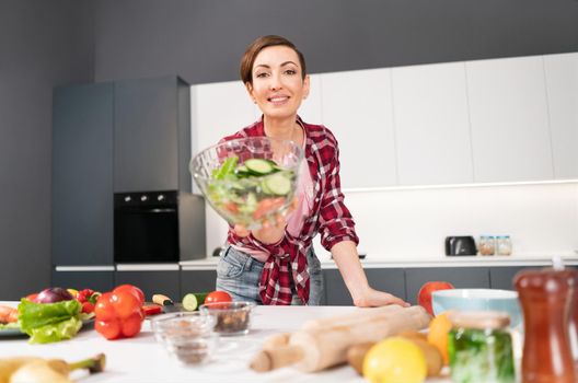 Happy young housewife holding bawl of salad showing it on camera prepared for a family dinner or girls night standing in the kitchen. Healthy food living. Healthy lifestyle.