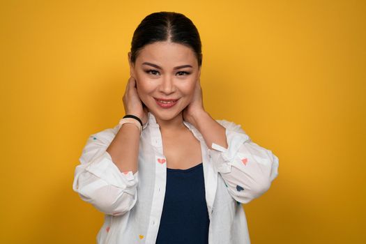 Cute mature Asian woman in a casual posing look on camera touching her back hair with both hands. isolated on yellow background. Human emotions, facial expression concept.
