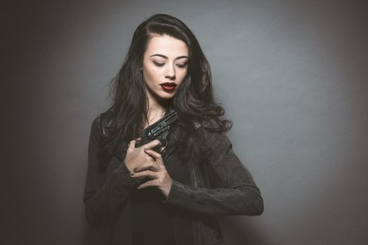 Secret agent beautiful young woman with red lips holding a gun wearing leather jacket. Dangerous young female model with professional make-up. Fashion, beauty, makeup, hairstyle and gun.