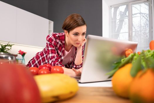 Watching interesting movie using laptop computer housewife forgot about cooking or baking stuck in a monitor. Young woman cooking in the kitchen. Healthy life living.