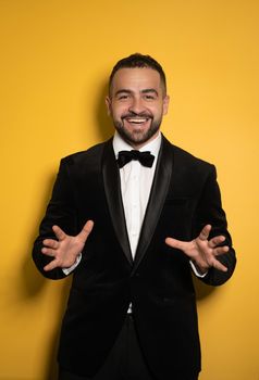 Good looking young businessman in black tuxedo smiling on camera, work profession lifestyle. Handsome young smiling caucasian man isolated on yellow background.