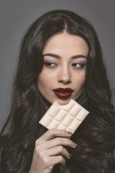 Sensual beautiful brunette young woman bite white bubbly chocolate wearing black jacket with beautiful long dark hair isolated on grey background. Young female model with professional make-up.