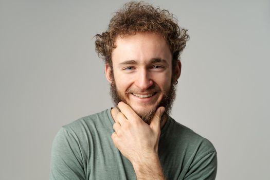 Handsome young guy posing in studio in olive t-shirt looking at camera touching his chin isolated on white background. Portrait of smiling young man with hands folded.