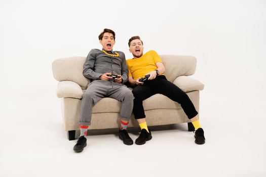 Friends playing video games while spend time together sitting on the small sofa looking at the camera isolated on white background. Group of guys sitting on couch and communicates on white.