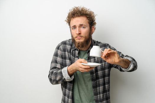 Good looking man with curly hair and beard holding inverted cup showing No coffee, guy wearing plaid long sleeve shirt isolated on white background. Crisis and lockdown concept.