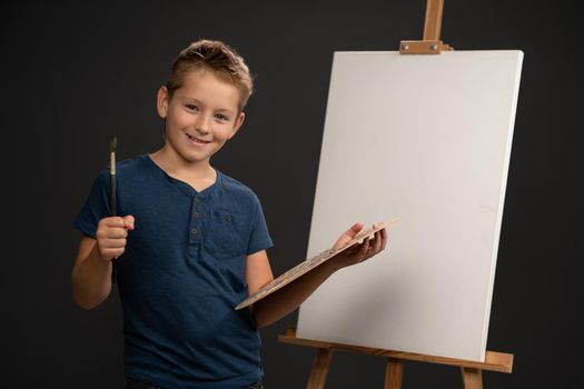 Charming boy in blue t-shirt looking at camera holding a palette with paints on the background of an easel with canvas. School of arts concept.