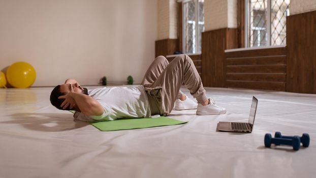 Young handsome bearded man doing abs crunches watching online work out videos on using laptop. Self motivated young man warming up doing special exercises for abs muscles.