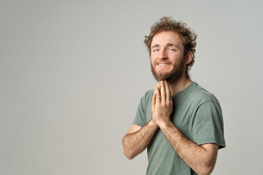 Begging for help put hands together as in prayer, handsome curly hair smiling man looking at camera tin need dressed in olive t-shirt isolated on white background.