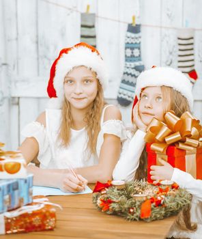 Two little girls sisters in santa hats are sitting at the table and composing a letter to santa claus, dreaming about gifts on christmas night, creating a wish list, The table is decorated with, .Christmas wreath, candles and full of Christmas gifts. White Wooden Wall with Christmas Stocking on it Background. Close-up . High quality photo