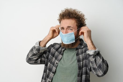 Disgustedly under duress puts on protective medical mask on his face to protect coronavirus, with curly hair, young man wearing plaid shirt and olive t-shirt under. White background.