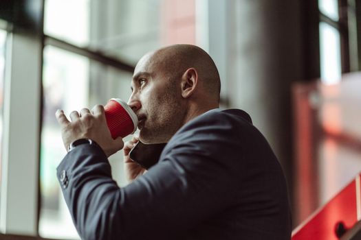 Mature business man in a business suit, talking on the phone while drinking coffee using disposable paper cup standing in a office building. Business concept.