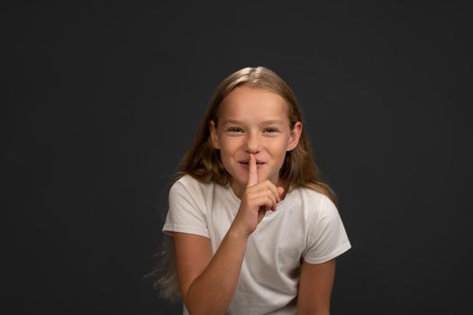Saying to keep silence little girl holds her finger on her lips telling everyone. Child wearing white t shirt smiling at the camera isolated on dark grey or black background.
