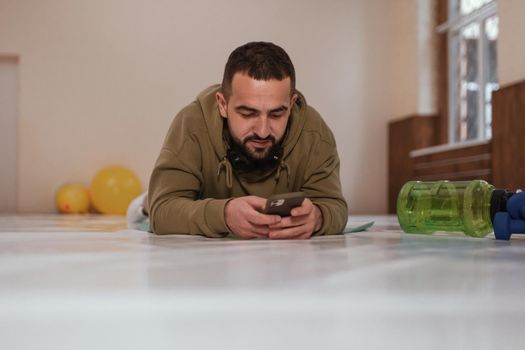 Young athletic man in a tracksuit, lies on the floor on a pad and watches video instructions or chatting on a smartphone. Video lessons on sports for those who are in quarantine.