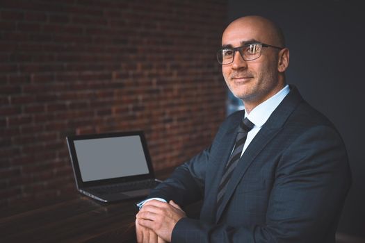 Mature bald businessman in eye glasses working alone at his desk in the office late at night, sitting half sideways with white screen on laptop and brick wall on the background.