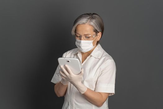 Doctor caucasian grey haired woman in protective mask work digital tablet gadget isolated on dark grey background.
