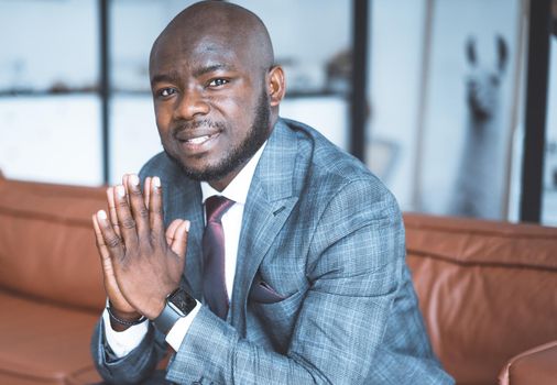 Young Nigerian Guy in a Formal Suit Sits with Folded Hands and Gazes into the Camera. Businessman from Africa is Ready to Provide his Conditions for Cooperation. Close-Up Portrait. Interior Background. High quality photo