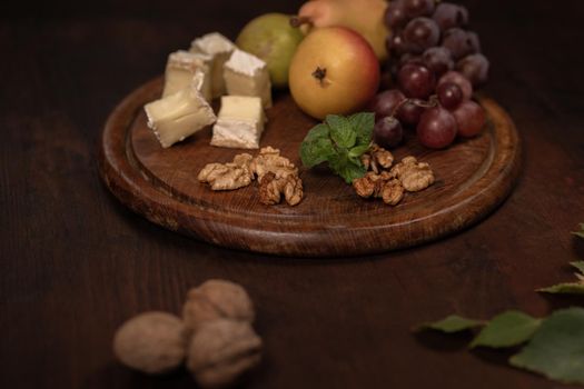 Brie cheese with fruits, grapes, walnuts on a dark wooden plate and wooden board. Wine snacks set: selection of cheese, grapes, pear and three walnuts on a wooden table. Side angle view.
