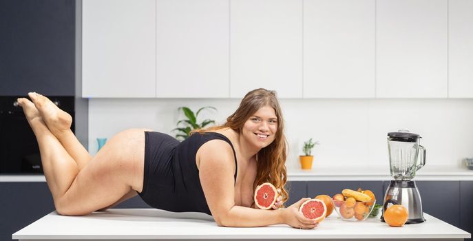 Sexy body positive chubby girl laying on the kitchen table wearing black swimsuit holding a fresh grapefruit in hands. Fat girl with long curly blond hair. Dieting and nutrition concept.