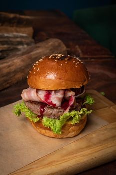 Craft burger with cherry sauce, fried slices of bacon, fresh lettuce next to the decorative wooden lodges. Dish served on a wooden cutting board and wood on background. Street food restaurant concept.