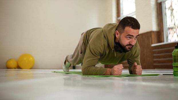 Young sports man doing elbow plank exercise in empty gym with yellow gym balls behind him all in khaki color track suit. Muscular sportsman doing exercises alone in gym.
