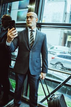 Confident businessman in suit reads information about in smartphone. Man holds mobile phone in front of him while standing next to window or elevator with busy street view in glassy wall.