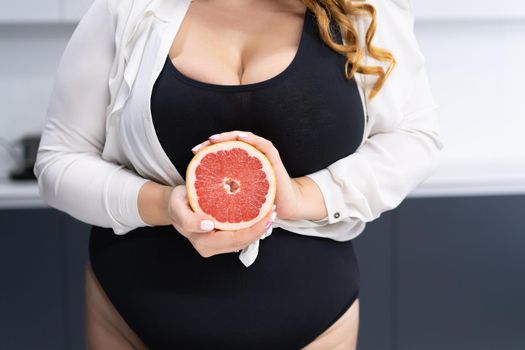 Close up curvy body young woman holding a cut half of a fresh grapefruit in hands standing on the modern kitchen at home with long blond hair. Dieting and nutrition concept.