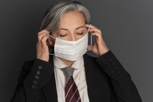 Grey-haired middle-aged business woman putting on a protective mask with eyes closed isolated on grey background. Portrait of modern senior woman in studio wearing business clothes.
