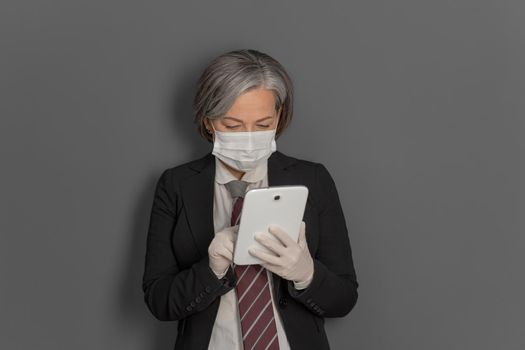 Caucasian businesswoman in protective mask work using digital tablet gadget standing next to the grey wall. Woman in black jacket on grey background. High quality photo.