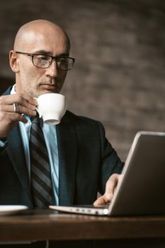 Drinking coffee mature business man working on laptop holding a cup of coffee sitting at table looking at laptop wearing eye glasses. Business concept.