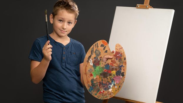 Holding a brush and palette with paints 10 year old boy in blue t-shirt looks at camera on the background of an easel with canvas. School of arts concept. Art concept.