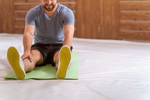 Young happy smiling fitness man doing stretching exercises at home. Amateur sports man in workout activity sitting on pad and stretch leaning to his toes. Hardworking guy exercising in apartment.