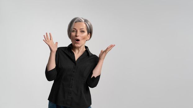 Grey haired mature woman doing a amazement gesture.Pretty mid aged grey haired woman in black shirt isolated on grey background. Human emotions, facial expression concept.