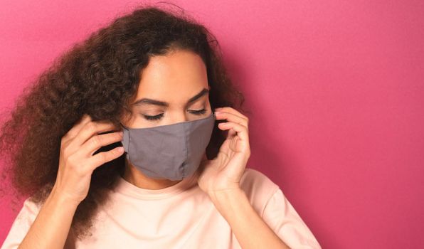 Young African American female in light peachy t-shirt putting on reusable face mask, to prevent others from corona COVID-19 and SARS cov 2 infection isolated on pink background.