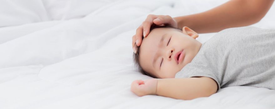 Young asian mother touch head of little baby girl with tender on bed in the bedroom, mom love newborn and care, woman with expression with child together, parent and daughter, family concept.