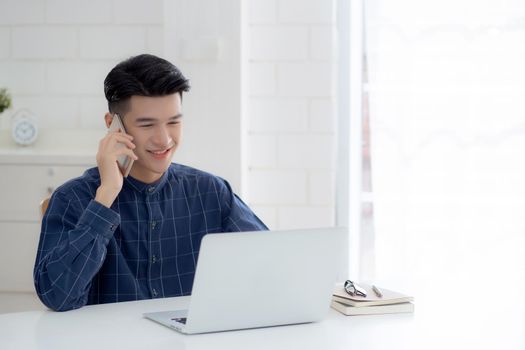 Young asian man talking phone and work from home with laptop computer, freelance using notebook to internet online and speak on smartphone, domestic life, business and communication concept.