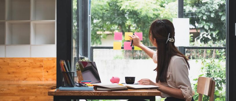 Business attractive young asian woman write notes on sticky note paper to arrange meeting with coworker sit in background take pictures through the glass, start up business and financial concept.