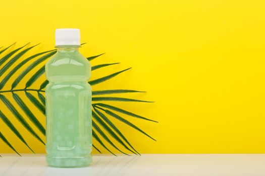 Detox drink or juice in transparent bottle with small pieces of fruits on white table against yellow background with palm leaf and copy space. Concept of healthy eating, dieting, weight loss and detox 