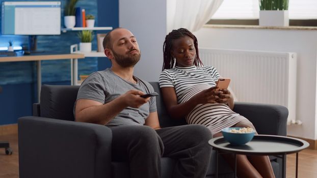 Young interracial couple expecting child and sitting on sofa at home. Caucasian man watching television and eating popcorn while african american pregnant woman using smartphone