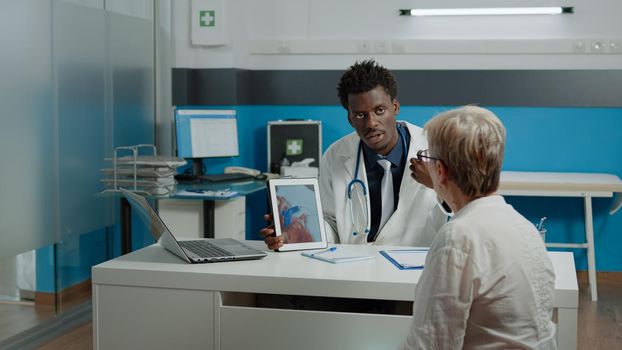 Healthcare specialist explaining cardiogram on tablet display to old patient with heart disease. Doctor showing cardiovascular system while holding device for anatomical checkup