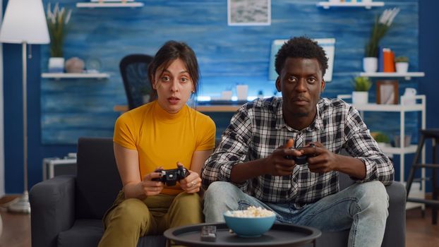 Modern interracial couple playing with joysticks for video game on console in living room. Young mixed race partners doing fun activity with television and controller together at home