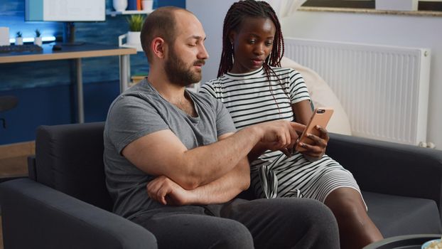 Young interracial couple looking at smartphone while black woman holding device at home. Mixed race husband and wife using technology and talking while sitting on couch in living room