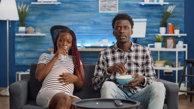 Black couple with pregnancy relaxing at home watching TV in living room. African american man bringing glass of water for pregnant woman eating popcorn on couch. Married people looking at camera