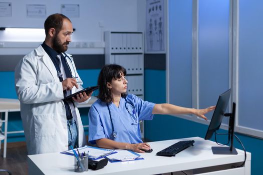 Team of medical specialists looking at computer screen and talking about healthcare system and treatment after hours. Doctor holding tablet for information while nurse pointing at monitor