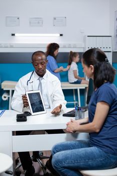 African american pediatrician doctor holding tablet computer explaining medical radiography to patient mother during clinical appointment in hospital office. Bones xray on screen. Health care service