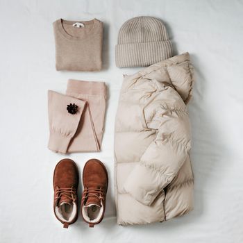 Women warm puffer light down jacket, beige cashmere sweater, pink knitted joggers, knitted bini hat, brown fur chukka suede boots on white bed sheet. Cozy winter clothing knolling in beige monochrome