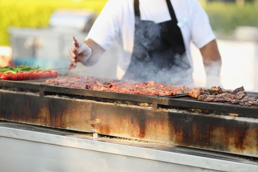 Close-up of professional male chef frying fresh meat on grill on open air. Man cook meal for visitors. Food, cafe, barbeque, cook, street kitchen concept