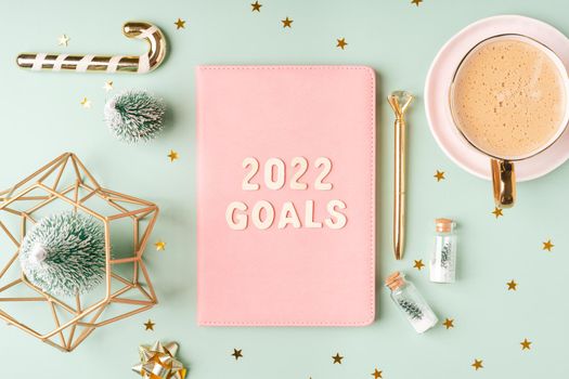 Top view Desktop Christmas pink notepad with 2022 goals letters text. Flat lay on green mint table background with planner, cup of coffee, candle, Christmas decoration, notebook and stationery.