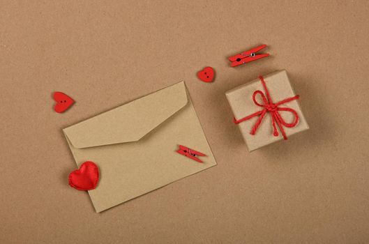 Close up Valentine gift box with red twine bow and card greeting envelope with wooden clothespins and hearts over brown paper background, flat lay, elevated top view, directly above